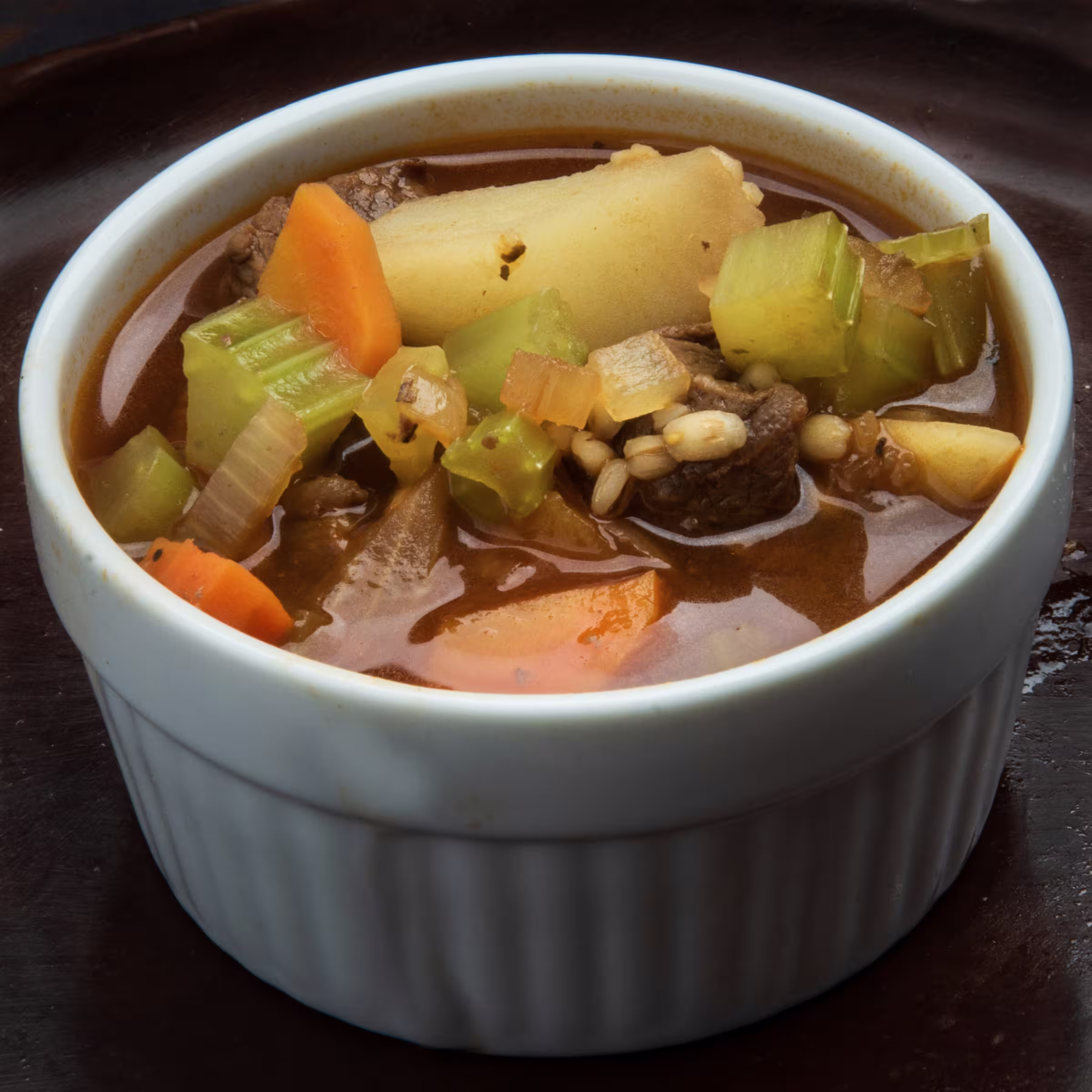 A white ceramic bowl of beef stew, with chunks of beef, fresh potatoes, carrots, and celery