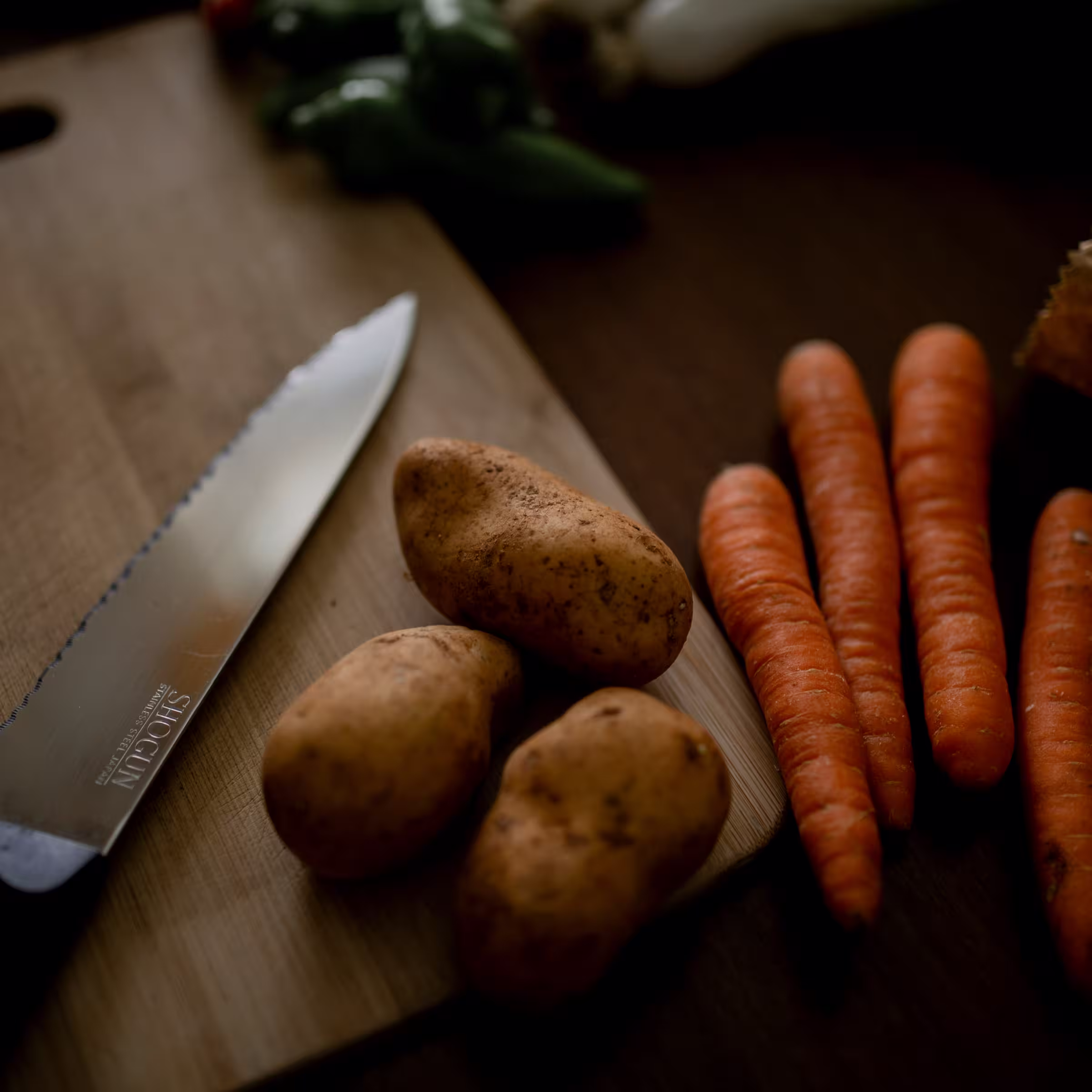 A wood cutting board with a knife and potatoes laying on it ready to cut and carrots off to the side.